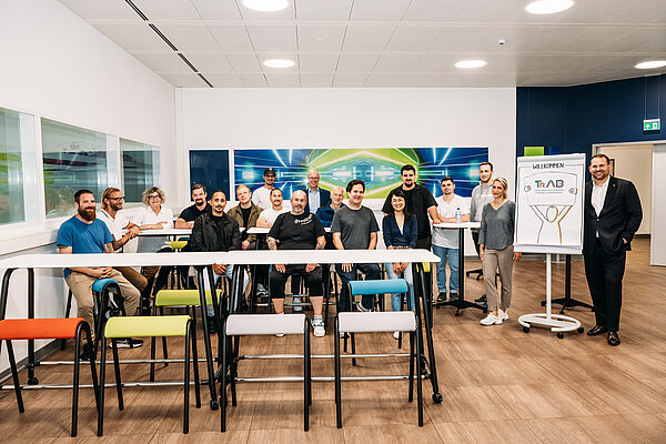 Teamfoto in einem modernen Besprechungsraum Gruppenfoto der Teilnehmenden des TrAB-Seminars mit Seminarleitung und Gästen bei Provadis. Die Gruppe sitzt und steht hinter Seminartischen. Rechts steht ein Flipchart mit der Aufschrift „Willkommen“ und dem TrAB-Logo.