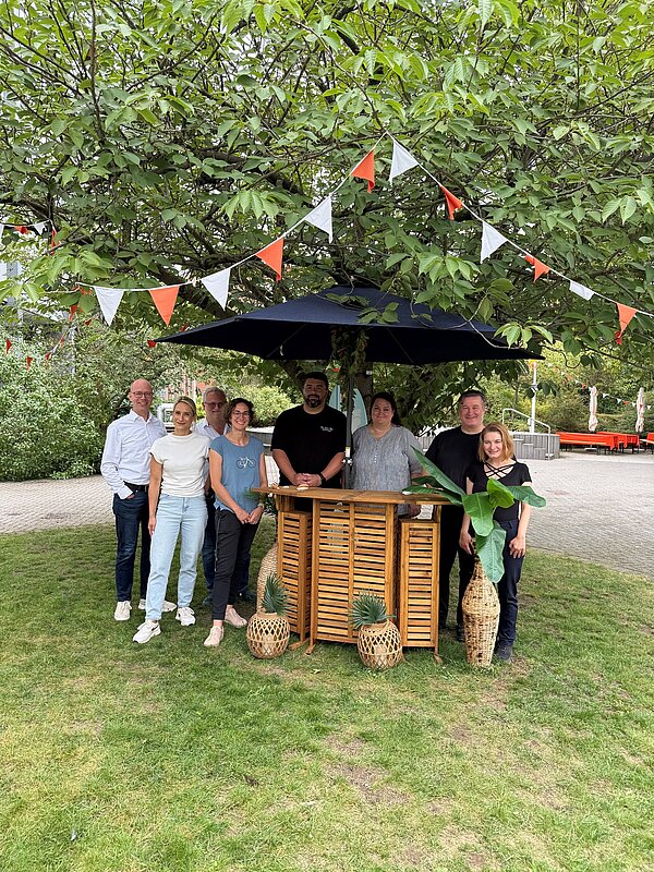 Mitarbeiterfoto vor einem Empfangsbereich im Freien. Acht Personen stehen nebeneinander im Innenhof des Provadis Campus und blicken in die Kamera