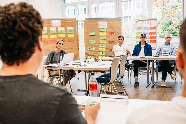 Teilnehmer eines Workshops mit Präsentationsmaterial Gesprächsrunde im TrAB-Seminar bei Provadis. Eine Frau sitzt mit einem Laptop gegenüber den Teilnehmenden. Im Hintergrund stehen Pinnwände und ein Flipchart mit bunten, beschrifteten Karten.