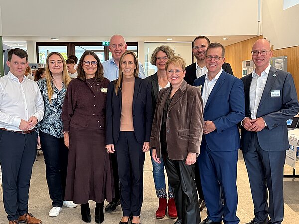 Gruppenfoto von Teilnehmern einer Veranstaltung. Gruppenfoto von zehn Teilnehmenden und Gästen der MINT-Messe 2026. Die Personen stehen nebeneinander im Eingangsbereich des Veranstaltungsgebäudes. Im Hintergrund sind weitere Messebesucher zu sehen.