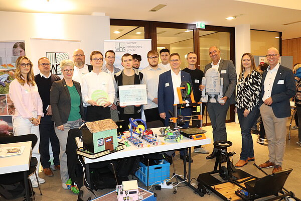 Gruppenfoto bei der 21. MINT-Messe: Mehrere Personen stehen hinter einem Tisch mit technischen Exponaten wie 3D-Druckern, Robotermodellen und einem Miniaturhaus. In der Mitte hält eine Person einen großen symbolischen Scheck, daneben eine runde Auszeichnung mit der Aufschrift ‚Provadis MINT 2025‘. Im Hintergrund sind Banner der Werner-Heisenberg-Schule zu sehen