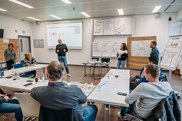 Teilnehmende der TrAB Inhouse-Schulung sitzen in einem Seminarraum in U-Form, während Provadis Geschäftsführer Holger Kison im Vordergrund steht und die Teilnehmenden begrüßt. Man sieht Inhalte zur pädagogischen Qualifizierung; Flipcharts und eine Präsentationsfolie verdeutlichen den praxisnahen Workshop-Charakter.