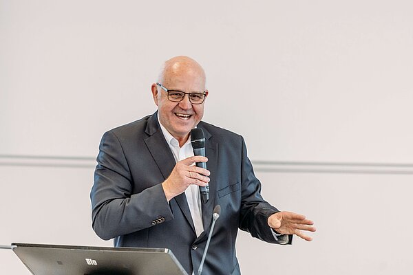 Abschiedsrede von Dr. Lemke Person in Business-Anzug spricht ins Mikrofon hinter einem Rednerpult, neutrale Wand im Hintergrund.