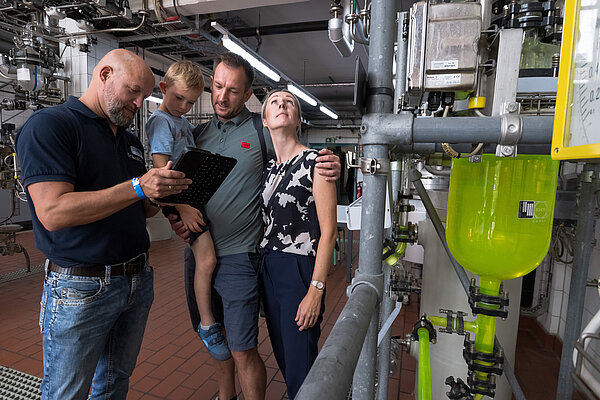 Familie und Fachkraft besprechen technische Details Eine Familie besichtigt gemeinsam mit einem Provadis-Mitarbeiter eine Produktionsanlage mit leuchtend grüner Flüssigkeit.
