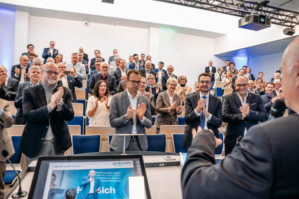 Standing Ovations Blick vom Rednerpult ins Auditorium, zahlreiche Personen stehen und applaudieren für Dr. Udo Lemke, Präsentationsfolie mit Provadis-Logo sichtbar.