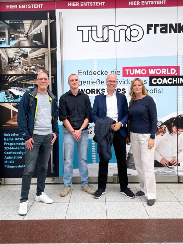 Vier Personen posieren vor den TUMO Frankfurt Werbetafeln, die den Start der neuen Kreativ- und Technologie-Workshops ankündigen. Neben ihnen sind Bilder der geplanten Innenräume und Workshop-Angebote zu sehen.