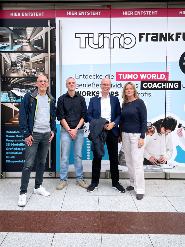 Vier Personen posieren vor den TUMO Frankfurt Werbetafeln, die den Start der neuen Kreativ- und Technologie-Workshops ankündigen. Neben ihnen sind Bilder der geplanten Innenräume und Workshop-Angebote zu sehen.