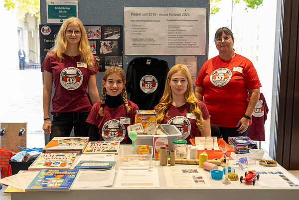 Vorstellung des Projekts EKS 2026 an einem Stand. Drei Schülerinnen und eine Lehrerin der Erich-Kästner-Schule Bürstadt stehen an einem Messestand. Auf dem Tisch sind zahlreiche Experimentiermaterialien, Bücher und selbst gebaute Modelle ausgestellt. Im Hintergrund informieren Plakate über das naturwissenschaftliche Schulprojekt.