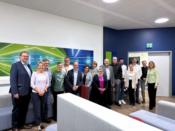 Runder Tisch Ausbildung 2025 Eine Gruppe von fünfzehn Personen steht gemeinsam in einem modernen Besprechungsraum bei Provadis. Die Gruppe lächelt in die Kamera, im Hintergrund ist eine bunte Wandgrafik mit grünen und blauen Lichtelementen zu sehen.