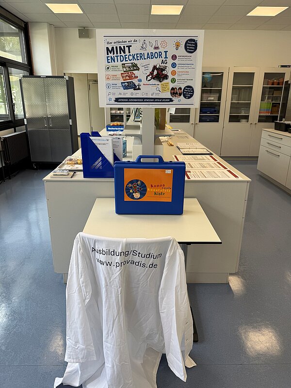 MINT Entdeckerlabor mit Lernmaterialien Ein ausgestatteter Naturwissenschaftsraum mit Experimentiermaterialien. Im Vordergrund stehen „Kunos coole Kunststoff-Kiste“ und ein weißer Laborkittel mit der Aufschrift „Ausbildung/Studium www.provadis.de“. Ein Plakat stellt die Angebote der „MINT Entdeckerlabor I AG“ in Biologie, Chemie, Physik und Technik vor.
