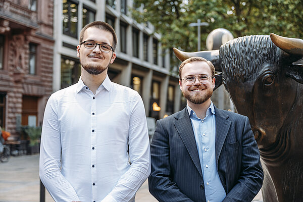 IHK Bestenehrung 2025 Die beiden ausgezeichneten Absolventen Johannes Kriehn (links) und Tommy Schilling (rechts) stehen nebeneinander vor dem Bullen-Denkmal an der Frankfurter Börse. Beide tragen Brillen und lächeln in die Kamera. Der Mann links trägt ein weißes Hemd, der Mann rechts ein hellblaues Hemd mit dunklem Anzug. Im Hintergrund sind historische und moderne Gebäude sowie Bäume zu sehen.