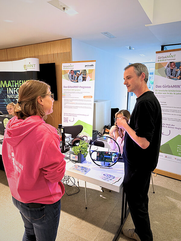 Zwei Personen stehen an einem Informationsstand mit Bannern zum Girls4MINT-Programm. Auf dem Tisch liegen Flyer, eine Pflanze und ein technisches Gerät mit Kabeln und Platine. Im Hintergrund sind Roll-Ups mit Texten zu MINT-Angeboten sichtbar.