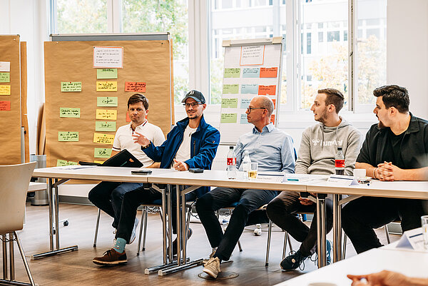 Teilnehmer bei einer Besprechung mit Notizen Fünf Teilnehmer des TrAB-Seminars sitzen an einer Tischreihe. Ein Mann mit blauer Jacke und Kappe gestikuliert im Gespräch. Hinter ihnen stehen Pinnwände mit beschrifteten Moderationskarten.